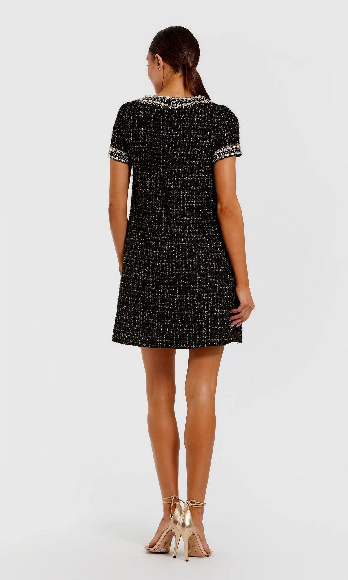 A woman stands facing away against a plain light background, wearing the Short Semi-Formal Dress: Mac Duggal 37075—a black short-sleeved boucle mini dress featuring silver trim and subtle rhinestone beading—paired with gold open-toe heels.